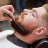 Young bearded man getting haircut by hairdresser while sitting in chair at barbershop. Barber soul.