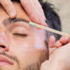 From above above crop woman using stick to apply wax on eyebrow of bearded young man with closed eyes during work in beauty studio